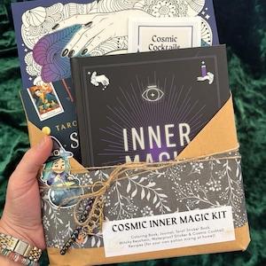 Puede incluir: Un kit Cosmic Inner Magic envuelto en papel floral gris, atado con hilo. El kit incluye un libro para colorear, un diario, un libro de pegatinas de tarot, un llavero de bruja y recetas de cócteles. El libro principal es negro con las palabras "INNER MAGIC".