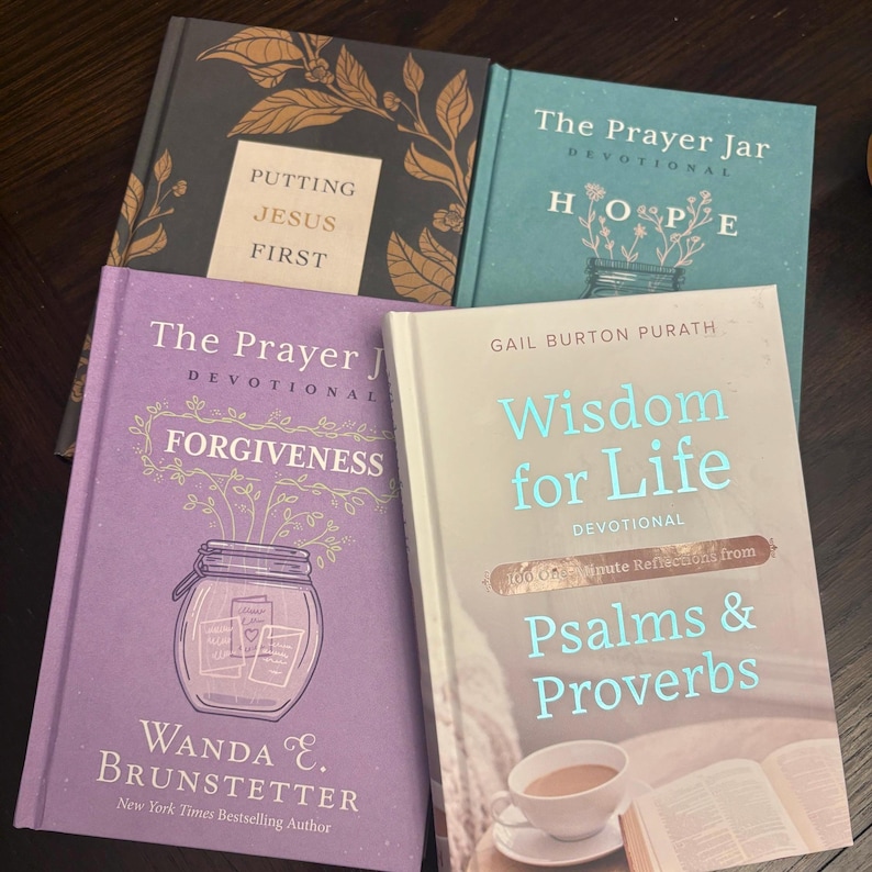 K&ouml;nnte beinhalten: Vier Hardcover-Andachtsb&uuml;cher mit Titeln wie "Putting Jesus First", "The Prayer Jar" und "Wisdom for Life". Die B&uuml;cher sind in verschiedenen Farben erh&auml;ltlich, darunter Teal, Lila und Schwarz, mit floralen und skriptartigen Designs. Die B&uuml;cher stammen von verschiedenen Autoren.