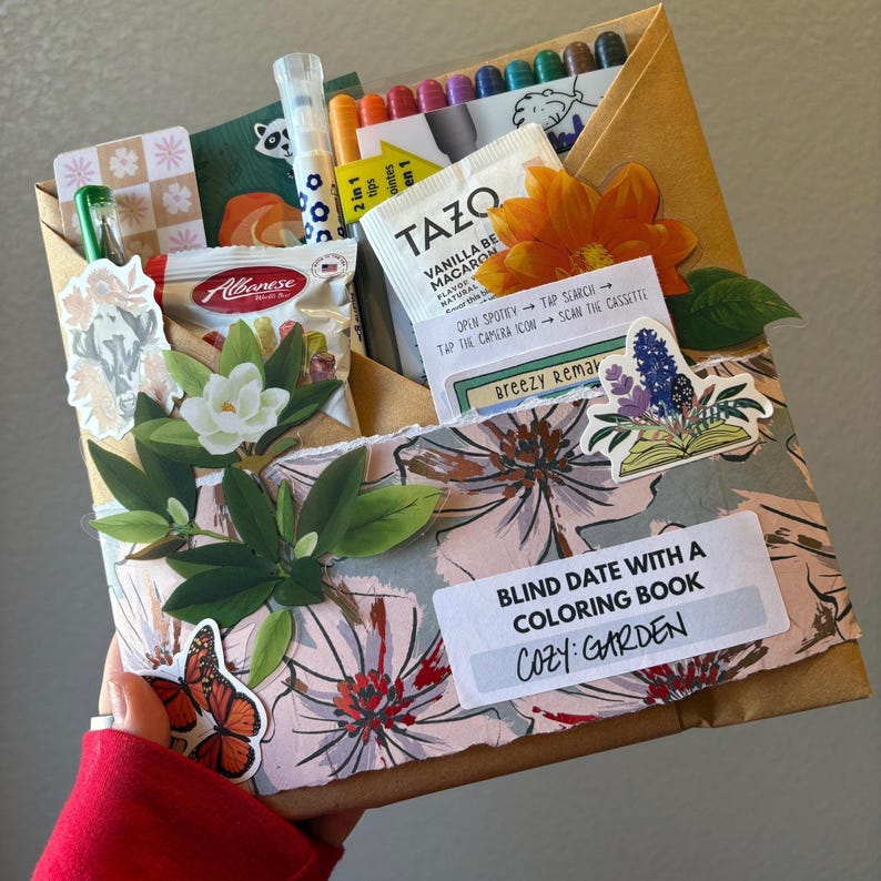 May include: A gift box labeled "Blind Date with a Coloring Book: Cozy Garden." The box is decorated with floral and leaf stickers, and contains coloring pens, tea, and snacks. The box is held by a person wearing a red sleeve.