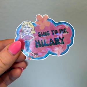 May include: A holographic sticker featuring a cartoon character with blonde hair holding a microphone, wearing a pink top and blue pants. The background is a pink cloud shape with orange flowers and the text "SING TO ME, HILARY" in dark green.