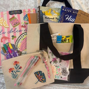 May include: A beige canvas tote bag with black handles containing a National Parks word search book, a KATEE ROBIT book, a reading list card, and a small pouch. The pouch features a floral patch, a leaf patch, and three pens with a beaded charm.