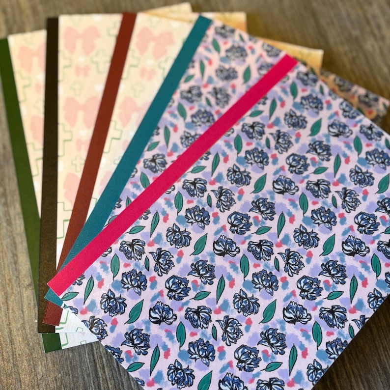 K&ouml;nnte beinhalten: Eine Sammlung von Notizb&uuml;chern mit gemusterten Einb&auml;nden. Die Einb&auml;nde zeigen Designs wie Blumenmuster, Schleifen und geometrische Formen. Jedes Notizbuch hat einen farbigen R&uuml;cken, mit Farben wie Rosa, T&uuml;rkis und Gr&uuml;n. Die Notizb&uuml;cher sind f&auml;cherf&ouml;rmig angeordnet.