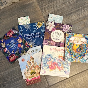 May include: A collection of coloring books with various themes, including butterflies, castles, mermaids, and floral designs. The books are arranged on a wooden surface, with titles like "Stress Free Coloring" and "Zen Patterns" visible.