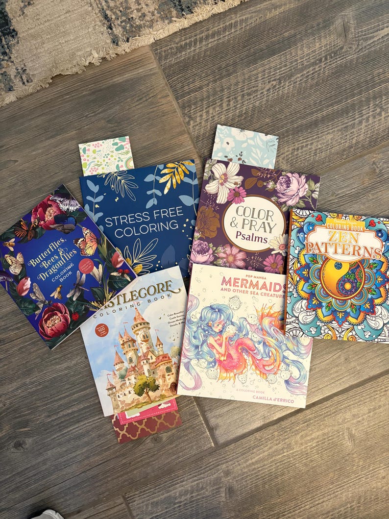 May include: A collection of coloring books with various themes, including butterflies, castles, mermaids, and floral designs. The books are arranged on a wooden surface, with titles like "Stress Free Coloring" and "Zen Patterns" visible.