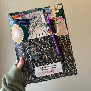 May include: A Halloween-themed gift box labeled "Blind Date with a Coloring Book Witchy." The box is decorated with stickers of ghosts, a witch, and a ghost with a bow. It contains coloring supplies and a coloring book.