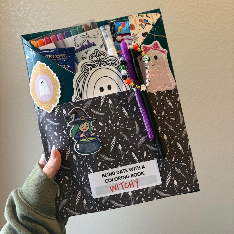 May include: A Halloween-themed gift box labeled "Blind Date with a Coloring Book Witchy." The box is decorated with stickers of ghosts, a witch, and a ghost with a bow. It contains coloring supplies and a coloring book.