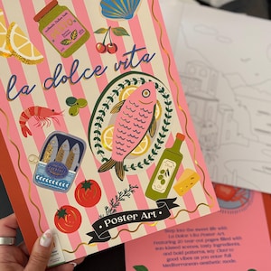 May include: A notebook with a pink and white striped cover featuring the words "la dolce vita." Illustrations of Italian food, including a pink fish, tomatoes, and sardines, are displayed. The words "Poster Art" are also visible.
