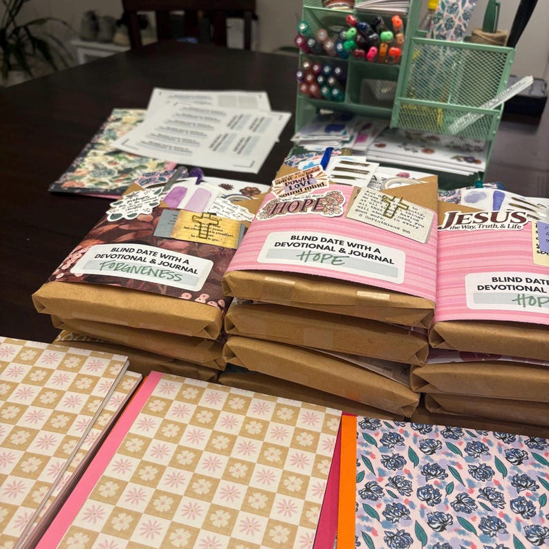 K&ouml;nnte beinhalten: Eine Sammlung von verpackten Andachtsjournalen mit dem Text "Blind Date with a Devotional & Journal" und den W&ouml;rtern "Forgiveness" und "Hope" auf den Etiketten. Die Journale sind auf einem Tisch mit anderen Schreibwaren gestapelt.