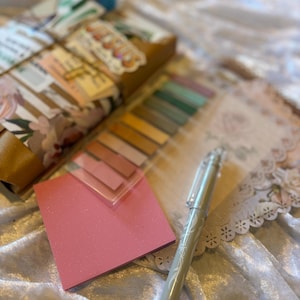 K&ouml;nnte beinhalten: Eine Sammlung von Schreibwaren, darunter ein pinkfarbener, glitzernder Haftnotizblock, ein durchsichtiger Kunststoffbeh&auml;lter mit bunten Haftf&auml;hnchen und ein silberner Stift. Ein verpacktes Paket mit Aufklebern und floralem Papier ist ebenfalls zu sehen.