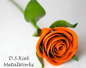 Puede incluir: Una sola rosa de metal naranja con hojas verdes. La rosa está hecha de metal y tiene un aspecto realista. D.S. Kiel Metalworks está impreso debajo de la rosa.