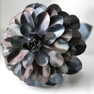 May include: A large, dark gray metal flower with a center made of smaller, curled metal pieces. The flower is made of many overlapping petals, giving it a layered appearance.