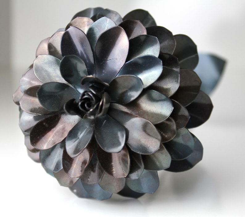 May include: A large, dark gray metal flower with a center made of smaller, curled metal pieces. The flower is made of many overlapping petals, giving it a layered appearance.