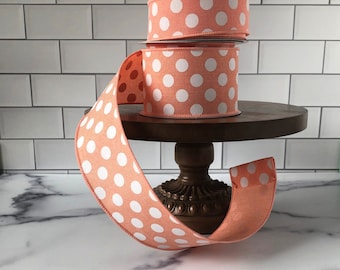 May include: A stack of three coral pink polka dot ribbon spools with white polka dots. The ribbon is displayed on a wooden cake stand.