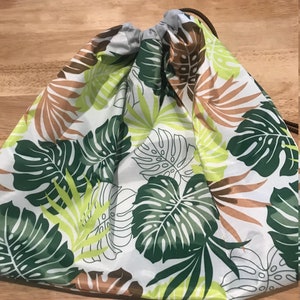 May include: A drawstring bag with a tropical leaf pattern. The bag features large green, light green, and brown leaves on a white background. The drawstring is brown.