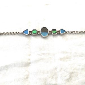 May include: Silver bracelet featuring a series of colorful, geometric-shaped stones. The bracelet has a chain link design with a toggle clasp. The stones include blue triangles, green squares, and an oval-shaped stone with blue and black hues.