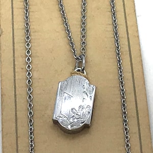 May include: A silver-toned pendant necklace with a detailed rectangular pendant. The pendant features an engraved design and hangs from a delicate chain. The necklace is displayed against a neutral background.