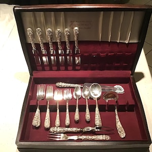 May include: A wooden silverware case with a red velvet interior holds an array of ornate silver utensils. The set includes knives, forks, spoons, and serving pieces, all with detailed floral designs on the handles. The case is open, revealing the complete set.
