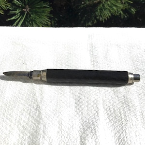 May include: A small, vintage pocket knife with a black handle and a silver blade. The knife is closed, with the blade tucked into the handle. The handle is cylindrical and appears to be made of wood or a similar material.