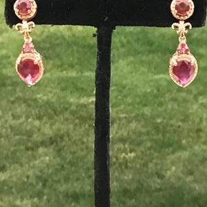 May include: Gold-toned drop earrings with a teardrop-shaped, deep pink gemstone. The earrings have a floral design and smaller pink stones. The background is a blurred green lawn.