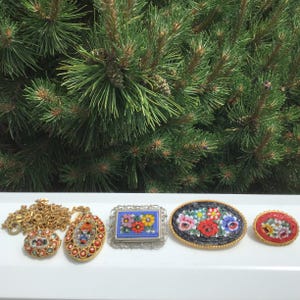 May include: A collection of vintage mosaic jewelry, including a necklace, pendants, and brooches. The pieces feature colorful floral designs and intricate detailing, set against a backdrop of evergreen foliage.