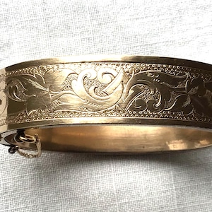 May include: A gold-toned hinged bangle bracelet featuring an intricate engraved floral and scrollwork pattern across the outer surface. The bracelet includes a secure side clasp mechanism and a small attached safety chain for added closure security.