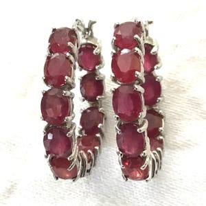 May include: A pair of hoop earrings featuring oval-cut, deep red gemstones set in a silver-toned metal. The gemstones are closely set, creating a continuous line around the hoop. The earrings are displayed against a white background.