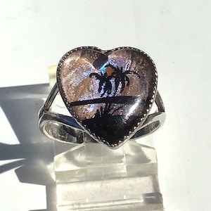 May include: Silver ring with a heart-shaped setting. The setting features a landscape design with palm trees and a sunset. The ring is displayed on a clear, rectangular stand.