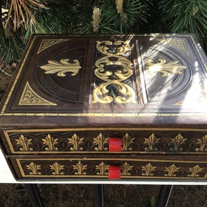 May include: A brown, antique-style box with gold accents and two drawers. The box features ornate gold leaf designs and red drawer pulls. The box is rectangular and appears to be made of leather or a similar material.