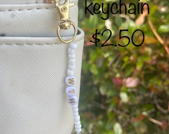 May include: White glass beaded keychain with the word "WITH" spelled out in gold letters. The keychain is attached to a gold metal clasp.