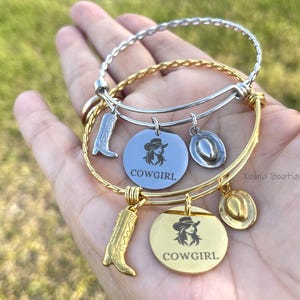 May include: A set of gold and silver-toned bangles with cowgirl-themed charms. The charms include a cowboy boot, a cowboy hat, and round pendants with a cowgirl silhouette and the word "COWGIRL".