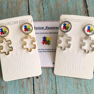 May include: A pair of gold and silver stainless steel earrings with puzzle piece charms. The charms feature a colorful puzzle piece design representing autism awareness. The earrings are displayed on white cards with the text "Stainless Steel" and "Xioma Boutique".