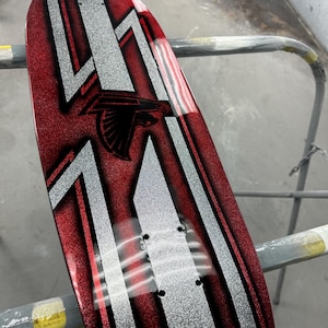 May include: A red and silver skateboard deck with a black Atlanta Falcons logo. The deck has a glitter finish and a black and silver striped design.