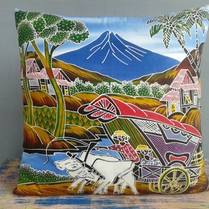 Op de afbeelding: Een kleurrijk kussenovertrek met een batik-ontwerp dat een landelijk tafereel voorstelt met een berg, huizen, palmbomen en een kar die door twee ossen wordt getrokken.
