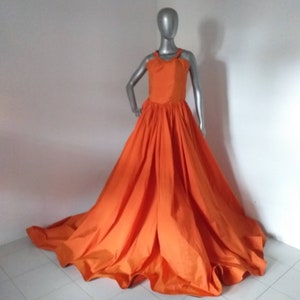 Könnte beinhalten: Ein orangefarbenes Abendkleid auf einer Schaufensterpuppe. Das Kleid hat einen Neckholder-Ausschnitt und einen weiten Rock, der in eine Schleppe übergeht. Der Stoff wirkt leicht und erzeugt einen voluminösen Effekt.