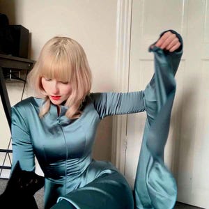 Könnte beinhalten: Türkisfarbener Satin-Jumpsuit mit langen Ärmeln und ausgestellten Beinen. Der Jumpsuit hat einen hohen Kragen und einen Reißverschluss vorne. Eine schwarze Katze ist im Bild zu sehen. Das Model hat blondes Haar und roten Lippenstift.