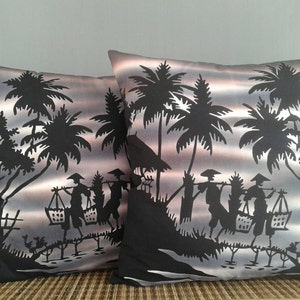 Op de afbeelding: Twee decoratieve kussens met een zwart-wit silhouetontwerp van palmbomen en mensen die manden dragen. De achtergrond is een verloop van grijs, roze en blauw.