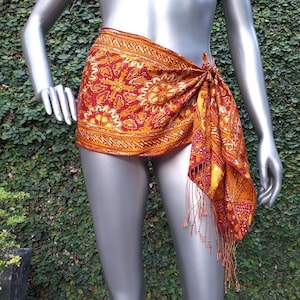 Op de afbeelding: Een rode en gouden sarong met franjes. De sarong is om de taille en heupen gebonden.