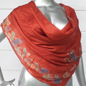 Puede incluir: Una bufanda de seda de color óxido con un patrón floral en tonos de azul, verde y rosa. La bufanda está drapeada sobre los hombros de un maniquí.