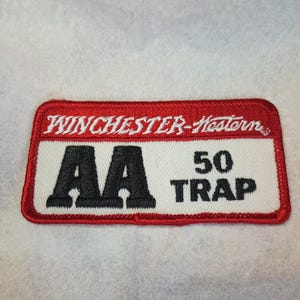 May include: A red and white embroidered patch with the text "Winchester-Western AA 50 Trap" in white.