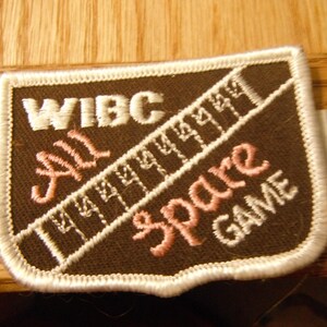May include: A brown and white embroidered patch with the text "WIBC All Spare Game" in white and pink lettering. The patch has a white border.