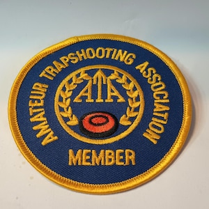 Può includere: Una toppa ricamata blu e oro con il testo "Amateur Trapshooting Association" e "Member". La toppa presenta un piattello di argilla stilizzato e le lettere "ATA" all'interno di una ghirlanda.