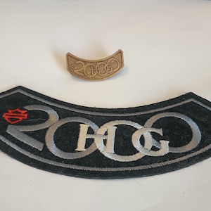 Puede incluir: Dos parches de Harley-Davidson. El parche más grande es negro con texto bordado plateado y un logotipo rojo. El texto dice "2000 H-D". También hay un pin más pequeño, de color marrón claro, con el texto "2000 H-D".