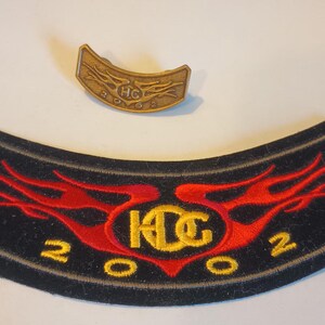 Puede incluir: Un parche bordado negro y rojo con el texto "K.D.C. 2002" en amarillo. El parche tiene un diseño de llamas y tiene forma de rocker de motocicleta. También se incluye un pequeño parche de metal dorado con el texto "H.D.C. 2002".