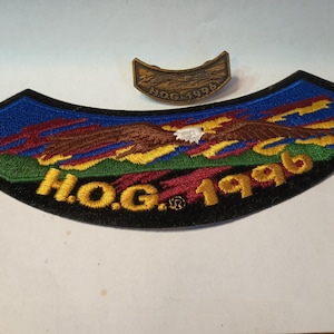 Può includere: Una toppa ricamata nera e dorata con un'aquila calva che vola sopra un paesaggio colorato. La toppa dice "H.O.G. 1996". È incluso anche un piccolo spillo di metallo dorato con il testo "H.O.G. 1996".