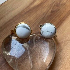 Könnte beinhalten: Zwei Ringe mit weißen Howlith-Steinen, die in Gold- und Silberfassungen gefasst sind. Die Ringe werden auf einem klaren herzförmigen Kristall präsentiert.