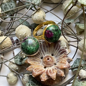 Puede incluir: Dos anillos de oro con engastes de piedras preciosas verdes y rojas. Los anillos están sobre un objeto de madera tallada en forma de flor. El objeto está rodeado de enredaderas marrones secas con hojas verdes.