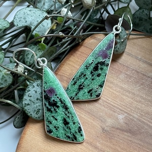 Op de afbeelding: Zilverkleurige oorbellen met een driehoekig design van natuursteen. De steen heeft een groene basis met zwarte en rode insluitsels, gezet in een zilveren montuur. De oorbellen worden gepresenteerd op een houten oppervlak, met groen op de achtergrond.