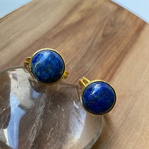 May include: Two gold rings with round blue lapis lazuli stones. The rings are on a wooden surface with a clear quartz heart.