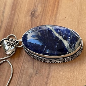 May include: Oval-shaped sodalite pendant with a silver-colored metal setting and chain. The deep blue stone has white veining. The pendant's setting has a decorative border and bail. The chain is a thin, silver-colored snake chain.