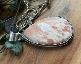 Può includere: Collana con catena d'argento e pendente a goccia. Il pendente presenta una pietra lucida nei toni del pesca, bianco e marrone rossiccio, incastonata in una lunetta d'argento. La collana è appoggiata su una superficie di legno.